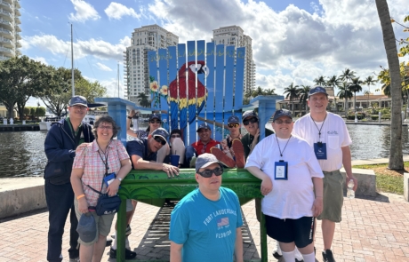 Travelers sitting and standing around an oversize decorated beach chair with a painted parrot on it.